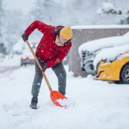 Man in red jacket shoveling snow during a heavy snowfall in a residential area.