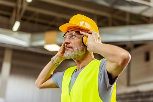 An industrial worker wears safety gear, including helmet and ear protection, in a brightly lit workspace, focusing on auditory safety.