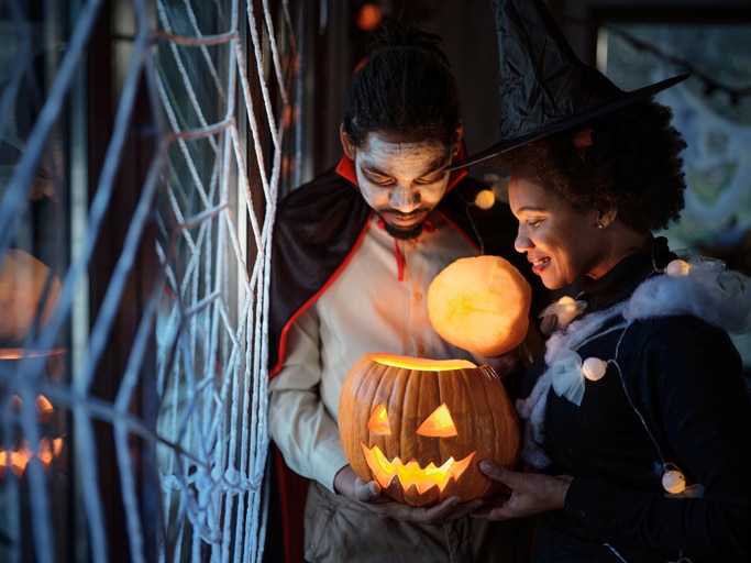 A couple dressed up for Halloween celebration looking inside of illuminated Jack O' Lantern at home.