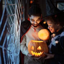 A couple dressed up for Halloween celebration looking inside of illuminated Jack O' Lantern at home.
